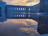 A rooftop scene with a reflective surface showing the mirrored image of a concrete wall with multiple rectangular windows. The sky is partly cloudy with a vibrant yellow and blue sunset, casting a serene glow. There is a horizontal wire with clothespins strung across the scene and a white water tank at the edge of the rooftop.