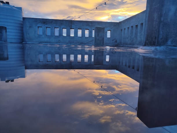 A rooftop scene with a reflective surface showing the mirrored image of a concrete wall with multiple rectangular windows. The sky is partly cloudy with a vibrant yellow and blue sunset, casting a serene glow. There is a horizontal wire with clothespins strung across the scene and a white water tank at the edge of the rooftop.