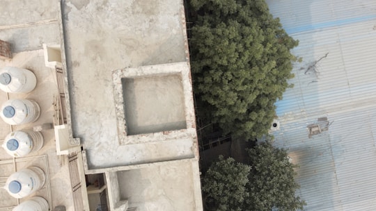An aerial view of a rooftop with several water tanks lined up along the left side. The roof has a rectangular opening in the center, while the right side features dense green foliage next to a large corrugated metal roof.