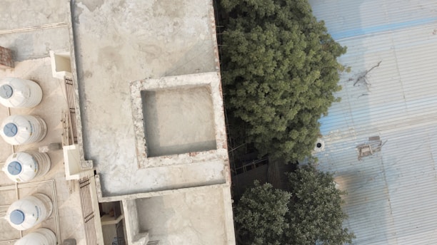 An aerial view of a rooftop with several water tanks lined up along the left side. The roof has a rectangular opening in the center, while the right side features dense green foliage next to a large corrugated metal roof.