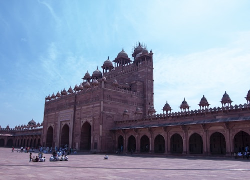 A large, intricately designed historical building made of red sandstone with multiple domes and arched entrances. The architecture features ornate detailing, typical of Mughal design. A spacious courtyard is populated with people sitting in groups, engaging in activities. The sky is clear and blue, providing a serene backdrop to the structure.