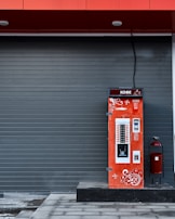A vibrant red coffee vending machine is stationed against a dark gray, paneled wall. The machine is adorned with white ornamental swirls and text in another language. A large cable extends from the top of the machine. To the right of the vending machine is a red, cylindrical trash can with a black stand.