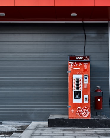 A vibrant red coffee vending machine is stationed against a dark gray, paneled wall. The machine is adorned with white ornamental swirls and text in another language. A large cable extends from the top of the machine. To the right of the vending machine is a red, cylindrical trash can with a black stand.