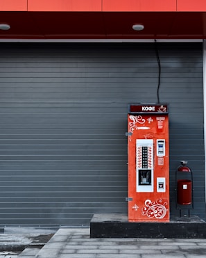 A vibrant red coffee vending machine is stationed against a dark gray, paneled wall. The machine is adorned with white ornamental swirls and text in another language. A large cable extends from the top of the machine. To the right of the vending machine is a red, cylindrical trash can with a black stand.
