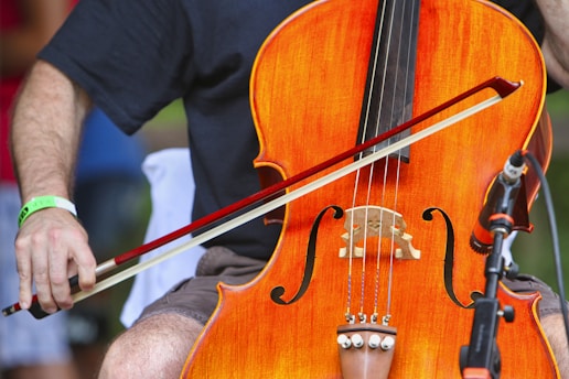 A close-up of a person playing a cello. The musician's left hand holds the neck of the cello while the right hand uses a bow to play. The cello is a bright orange-brown color and stands out prominently. A microphone is positioned near the instrument, suggesting a performance setting. The musician wears a black shirt and shorts, and a green wristband is visible.