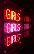 A neon sign displaying the word 'GIRLS' three times in bright pink and red colors, mounted on a dark background, creating a vibrant and eye-catching effect.