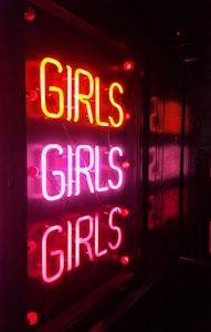 A neon sign displaying the word 'GIRLS' three times in bright pink and red colors, mounted on a dark background, creating a vibrant and eye-catching effect.