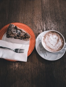 A slice of moist, homemade cake on a delicate ceramic plate with a cup of cappuccino.