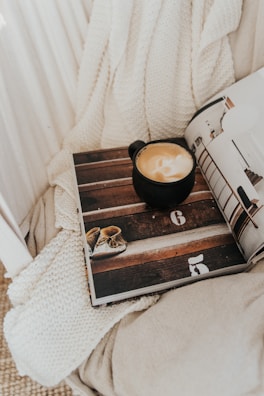 An artistic shot of a coffee tumbler next to a cozy knitted blanket and an open book.