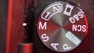 Close-up view of a camera mode dial on a digital camera with various settings including AUTO, P, S, M, and scene modes indicated in white lettering on a textured black surface. The edge of the dial has a metallic sheen reflecting red lighting.