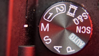 Close-up view of a camera mode dial on a digital camera with various settings including AUTO, P, S, M, and scene modes indicated in white lettering on a textured black surface. The edge of the dial has a metallic sheen reflecting red lighting.