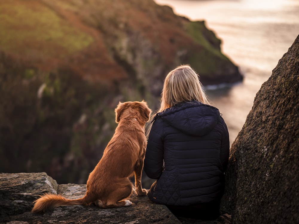 Donna in giacca nera che si siede accanto al cane marrone sulla roccia vicino allo specchio d'acqua durante il giorno