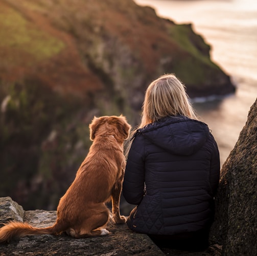 woman in black jacket sitting beside brown dog on rock near body of water during daytime