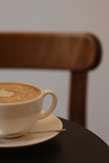 A creamy latte served in a white cup and saucer sits on a dark table. The background features a blurred wooden chair, adding a warm and cozy ambiance.