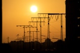 A silhouetted view of power lines and towers against a vibrant orange sunset or sunrise sky. The sun is positioned prominently on the left, casting a warm glow over the scene. The image captures the stark contrast between the dark outlines of the towers and the bright sky.