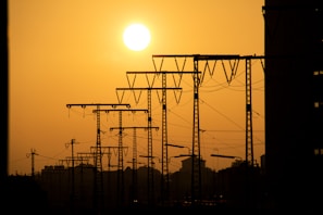 A silhouetted view of power lines and towers against a vibrant orange sunset or sunrise sky. The sun is positioned prominently on the left, casting a warm glow over the scene. The image captures the stark contrast between the dark outlines of the towers and the bright sky.