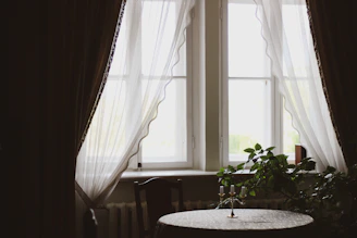 A serene corner with soft natural light filtering through sheer curtains onto a simple wooden table adorned with a small potted plant and a lit candle.