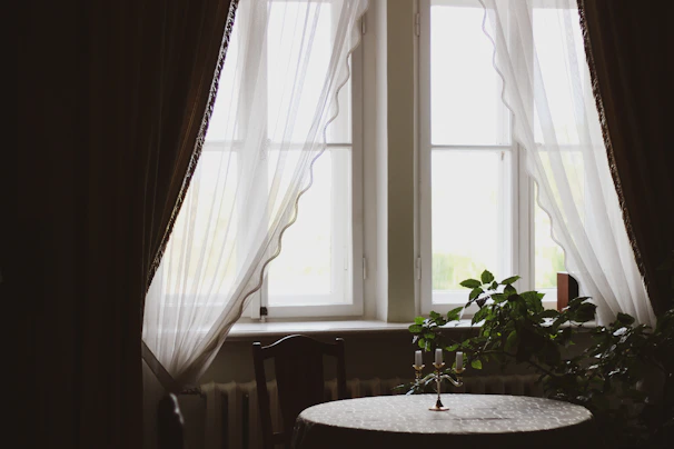 A serene corner with soft natural light filtering through sheer curtains onto a simple wooden table adorned with a small potted plant and a lit candle.