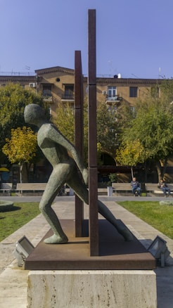 A modern sculpture of a human figure in a kinetic pose, seemingly pushing against or stepping through vertical metal beams. The sculpture is situated in an outdoor area with green grass, concrete pathways, and trees with autumn-colored leaves. There are low-rise buildings with balconies in the background. A couple of people are sitting on benches nearby, enjoying the sunny weather.