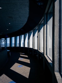 A curved interior corridor with large vertical windows that allow sunlight to cast geometric shadows on the floor. The design incorporates a mix of concrete surfaces and a modern architectural aesthetic, while blue markers on the floor suggest social distancing measures.
