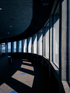 A curved interior corridor with large vertical windows that allow sunlight to cast geometric shadows on the floor. The design incorporates a mix of concrete surfaces and a modern architectural aesthetic, while blue markers on the floor suggest social distancing measures.