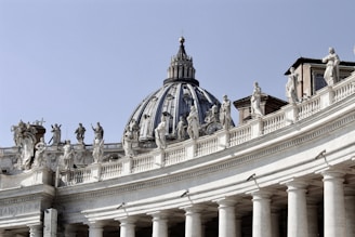 A large architectural structure featuring a classic dome and ornate statues lining the top of an arched colonnade. Each statue is distinct, and the building exhibits intricate details typical of Baroque architecture.