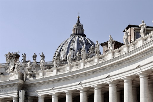 A large architectural structure featuring a classic dome and ornate statues lining the top of an arched colonnade. Each statue is distinct, and the building exhibits intricate details typical of Baroque architecture.