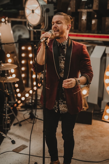 A man is singing into a microphone on a stage, surrounded by vintage marquee lights and musical equipment. He is wearing a dark patterned shirt and a red velvet jacket with black lapels.