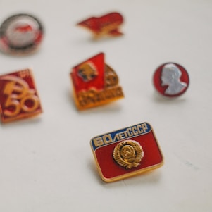 A collection of Soviet-era pins arranged on a light background. The pins are of various designs, featuring symbols and text associated with Soviet motifs, including the hammer and sickle. The badges display a combination of red, gold, and silver tones, indicative of the USSR's emblematic colors.