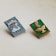 Two rectangular metal pins are placed on a light surface. The left pin features a blue background with a white swan and a castle, and text in Cyrillic script. The right pin has a green background with a golden ship and Cyrillic text. Both pins exhibit intricate designs and vibrant colors, suggesting a cultural or historical theme.