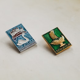 Two rectangular metal pins are placed on a light surface. The left pin features a blue background with a white swan and a castle, and text in Cyrillic script. The right pin has a green background with a golden ship and Cyrillic text. Both pins exhibit intricate designs and vibrant colors, suggesting a cultural or historical theme.