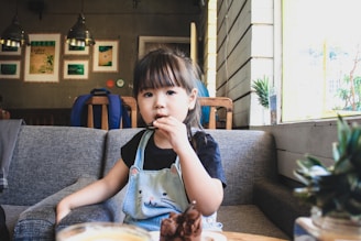 A young child is sitting on a grey couch in a cozy café setting, holding a cookie. The child is wearing a blue overall with a cat design on the front and a black t-shirt underneath. The background features a wooden paneled wall, framed pictures, hanging lights, and some potted plants on the window sill.