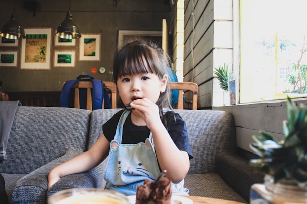 A young child is sitting on a grey couch in a cozy café setting, holding a cookie. The child is wearing a blue overall with a cat design on the front and a black t-shirt underneath. The background features a wooden paneled wall, framed pictures, hanging lights, and some potted plants on the window sill.