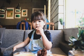 A young child is sitting on a grey couch in a cozy caf&eacute; setting, holding a cookie. The child is wearing a blue overall with a cat design on the front and a black t-shirt underneath. The background features a wooden paneled wall, framed pictures, hanging lights, and some potted plants on the window sill.