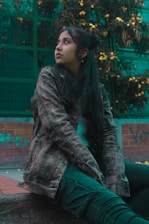 A young person sits on a ledge, looking away thoughtfully. They are wearing a denim jacket and dark pants, with their hair tied up. Behind them are green metal fences covered with dense foliage and bright yellow flowers.