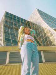 A young person stands confidently in front of a modern building with a unique curved glass facade. They are wearing a colorful jacket, a white crop top, and blue jeans. The perspective is from below, making the person look powerful against the urban architectural background.