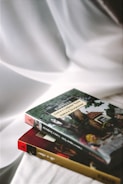 Two books are placed on a smooth white fabric surface. The book on the top features a cover with an illustration of a classic story involving characters and a scenic backdrop. The book underneath has a maroon and gold spine.