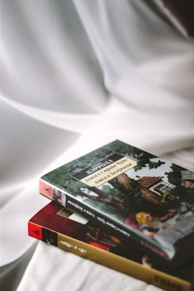 Two books are placed on a smooth white fabric surface. The book on the top features a cover with an illustration of a classic story involving characters and a scenic backdrop. The book underneath has a maroon and gold spine.
