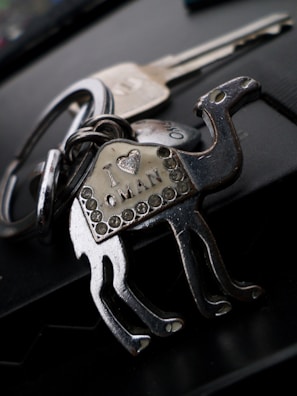 A metallic keychain shaped like a camel with decorative rhinestones. The body of the camel has an inscription that reads 'I ♥ Oman'. In the background, there is a blurred view of two metal keys attached to the keychain.
