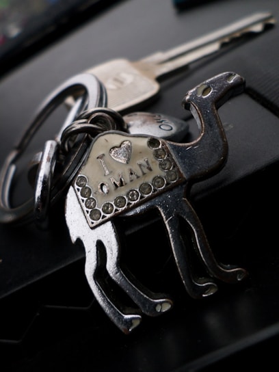 A metallic keychain shaped like a camel with decorative rhinestones. The body of the camel has an inscription that reads 'I ♥ Oman'. In the background, there is a blurred view of two metal keys attached to the keychain.