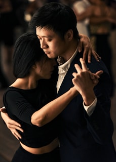 A couple is engaged in a close, intimate dance, with their eyes closed and arms wrapped around each other. They are dressed elegantly, and the background appears to be dimly lit, suggesting a moody, romantic atmosphere.