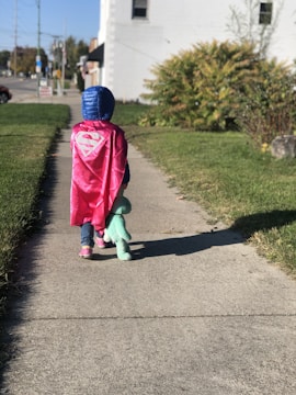 A child dressed in a pink superhero cape and a blue hooded jacket is walking on a sidewalk, carrying a green stuffed dinosaur toy. The scene is set in a suburban area with green grass, bushes, and a white building in the background under a clear sky.