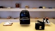A stylish display in a store showcasing a variety of footwear and accessories. In the center, there is a black leather backpack with a prominent brand logo. Below, a black woven handbag with a gold 'P' detail. Surrounding the central items, various pairs of shoes, including sandals, high heels, and sneakers, are neatly arranged on shelves.
