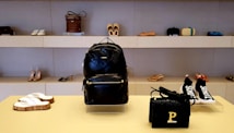 A stylish display in a store showcasing a variety of footwear and accessories. In the center, there is a black leather backpack with a prominent brand logo. Below, a black woven handbag with a gold 'P' detail. Surrounding the central items, various pairs of shoes, including sandals, high heels, and sneakers, are neatly arranged on shelves.