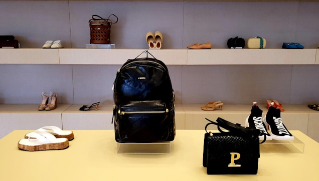 A stylish display in a store showcasing a variety of footwear and accessories. In the center, there is a black leather backpack with a prominent brand logo. Below, a black woven handbag with a gold 'P' detail. Surrounding the central items, various pairs of shoes, including sandals, high heels, and sneakers, are neatly arranged on shelves.