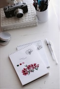 A vintage camera sits on a decorative box, alongside a white cup filled with pens and pencils. Below, a spiral-bound notebook features floral sketches and color swatches in red and green hues. A clip holds a pen on another pad containing dotted paper.