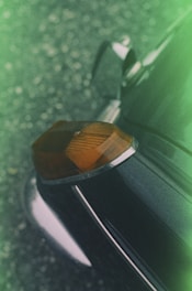 A close-up angle of a vintage car focuses on the rear corner, showcasing an orange tail light with chrome accents against a dark, slightly reflective car body. A soft, green gradient frames the image, adding an abstract, artistic element to the scene.