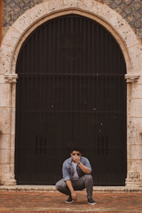 A person wearing sunglasses is crouched down, covering part of their face with their hand, in front of a large, arched, black iron gate that is set within a stone archway. The archway is decorated with an ornate tiled pattern above and around it.