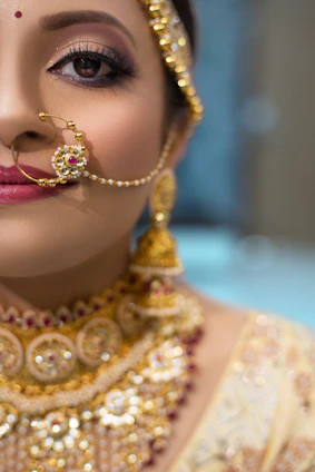 A close-up of a person wearing intricate and ornate traditional jewelry. The focus is on an elaborate gold nose ring and matching earrings with detailed craftsmanship. The makeup is elegant, with emphasis on the eyes and lips, adding a touch of sophistication and cultural richness.