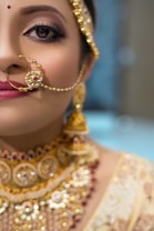 A close-up of a person wearing intricate and ornate traditional jewelry. The focus is on an elaborate gold nose ring and matching earrings with detailed craftsmanship. The makeup is elegant, with emphasis on the eyes and lips, adding a touch of sophistication and cultural richness.